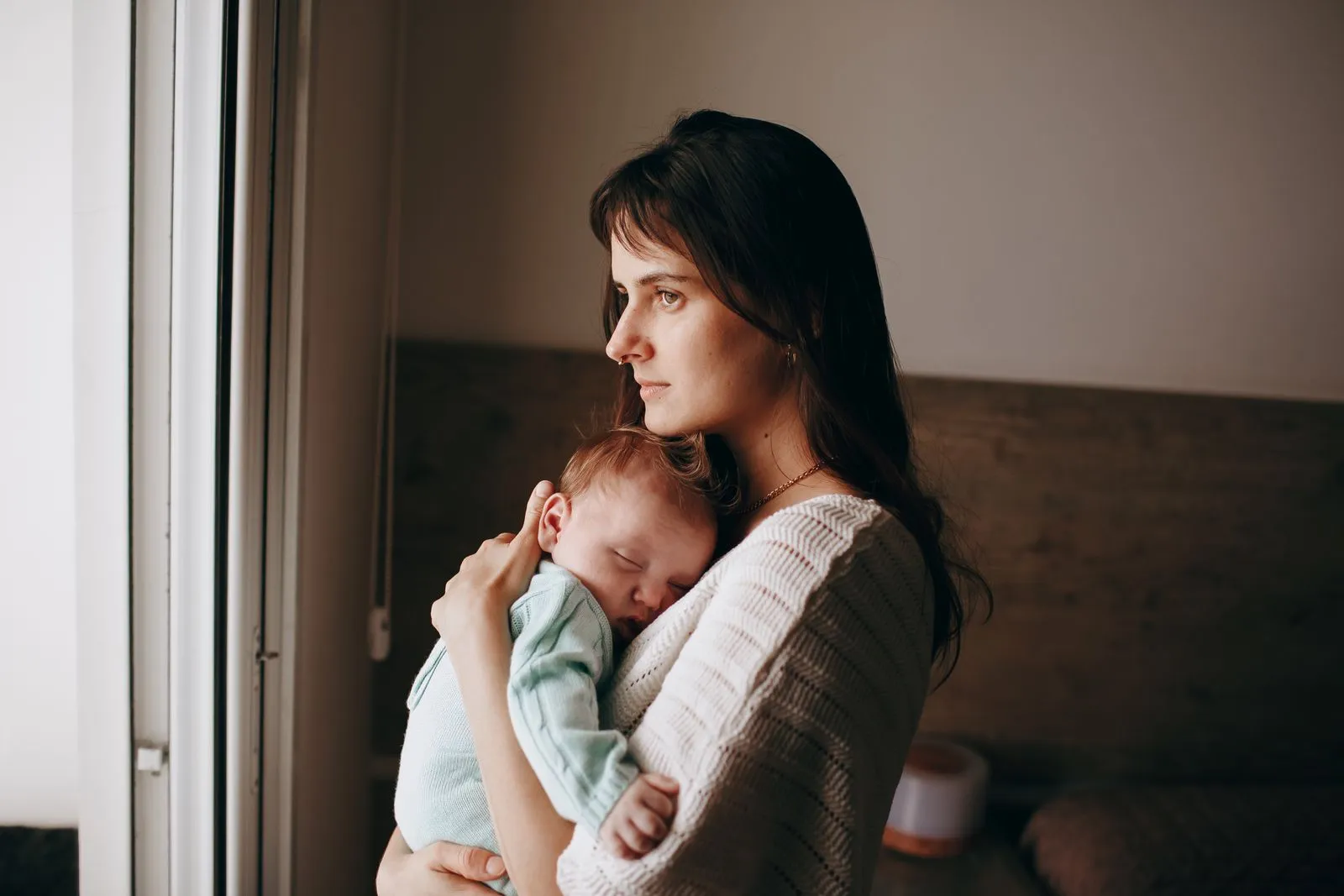 Mãe segurando o bebê recém-nascido adormecido no colo, perto de uma janela